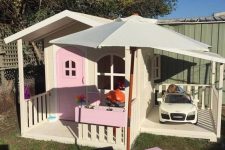 a pretty pink and neutral kids’ playhouse with a pink door, potted plants, a little dining set with an umbrella and a car