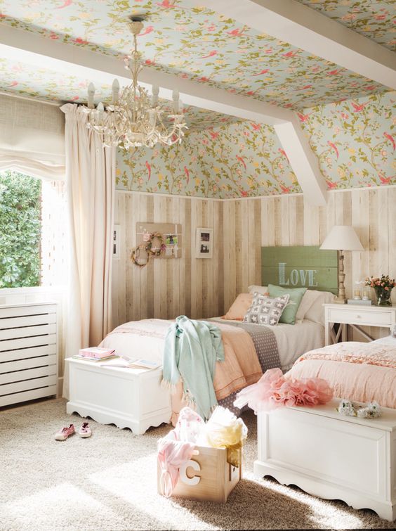 a shabby chic kids' room with floral wallpaper and wood paneling, white vintage furniture, pastel bedding and a chic chandelier