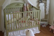 a shabby chic kid’s room with light green walls, a green crib with a canopy, floral bedding, a ladder for storage, fabric flowers and vintage toys