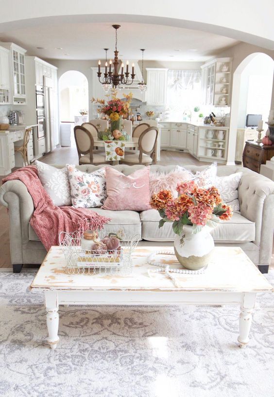 a shabby chic living room with a grey sofa, a white shabby table, pink pillows and blankets and blooms