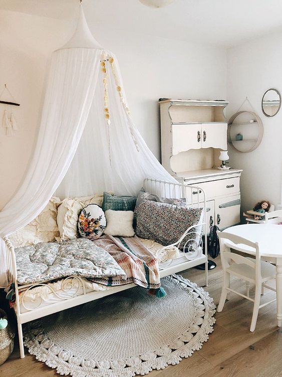 a shabby chic meets boho kid's room with neutral walls, white vintage furniture, pretty bright bedding and pillows and a canopy over the bed