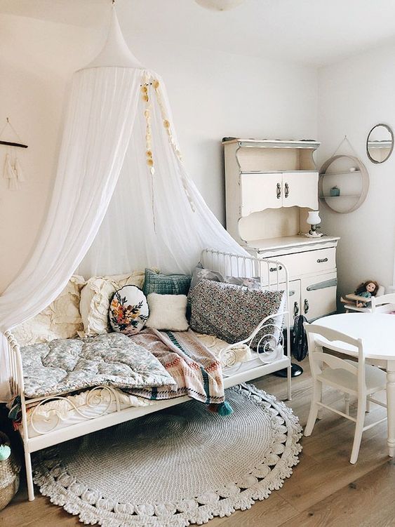 a shabby chic meets boho kid's room with neutral walls, white vintage furniture, pretty bright bedding and pillows and a canopy over the bed