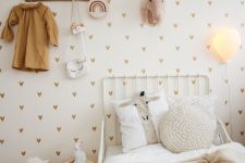 a stylish neutral Scandi kid’s room with a heart printed wall, a white metal bed with neutral bedding, a small nightstand and a rack plus some decor
