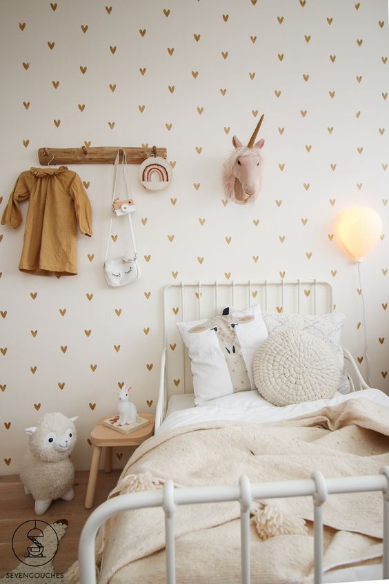 a stylish neutral Scandi kid's room with a heart printed wall, a white metal bed with neutral bedding, a small nightstand and a rack plus some decor