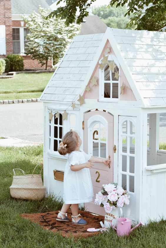 a tiny and cute white and pink kids' playhouse with a pink door, a star garland and blooms is ideal for a little kid