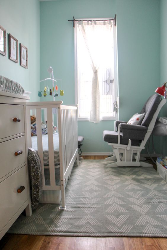 a tiny modern nrusery with light blue walls, a white crib and a dresser, a grey chair, a gallery wall and printed textiles