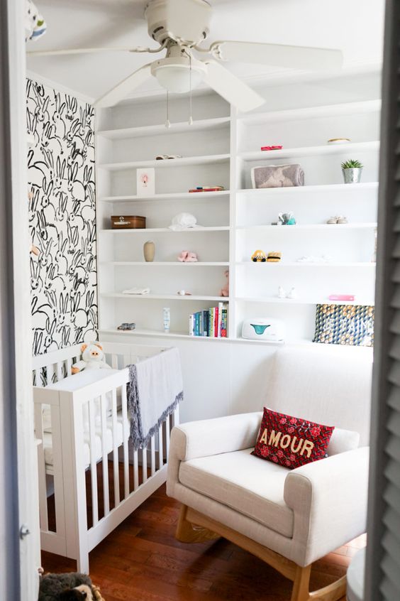 a tiny neutral nursery with a bunny accent wall, built-in shelves, a white crib and a rocker plus some bright textiles
