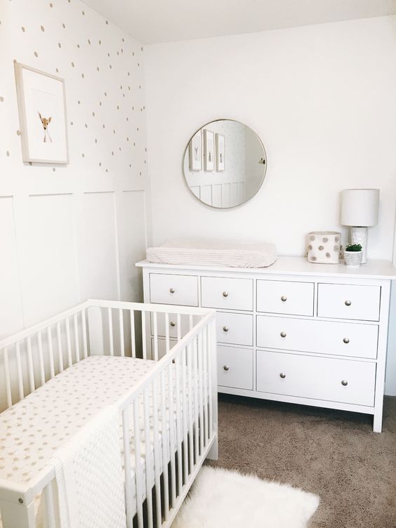 a tiny neutral nursery with a white crib and a dresser, layered rugs, a polka dot accent wall and a round mirror