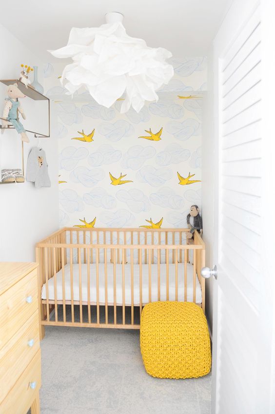 a tiny nursery with an accent wall, a stained crib with neutral bedding, a stained dresser, a yellow pouf and some shelves