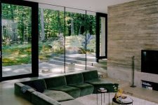 a welcoming modern conversation pit done with a TV, a green sofa, black round tables and a gorgeous view of the forest through a glass wall