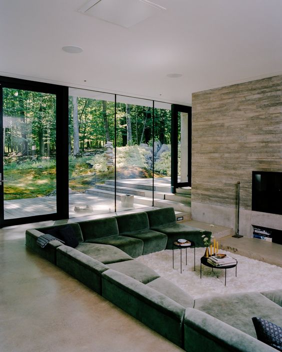 a welcoming modern conversation pit done with a TV, a green sofa, black round tables and a gorgeous view of the forest through a glass wall