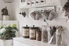 a white shabby chic kitchen with vintage furniture, a cabinet with a curtain, potted blooms, a metal pendant lamp