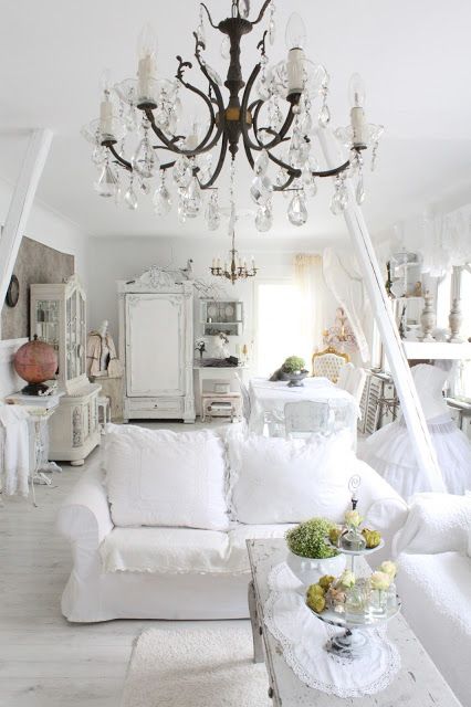 a white shabby chic living room with sofas, a crystal chandelier and a dining zone a bit further done with a cohesive look