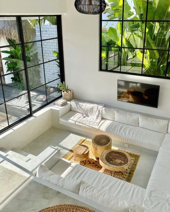 an all-white conversation pit with a built-in sofa with white upholstery, wood coffee tables and a bold boho rug plus large windows for more natural light