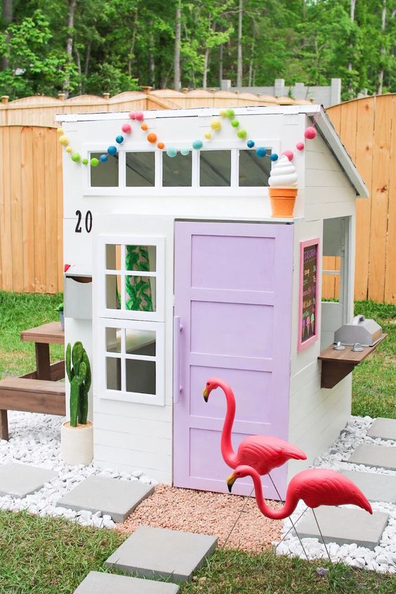 an ice cream inspired kids' playhouse with a lilac door, a colorful pompom garland, a cactus and a stained dining set behind it