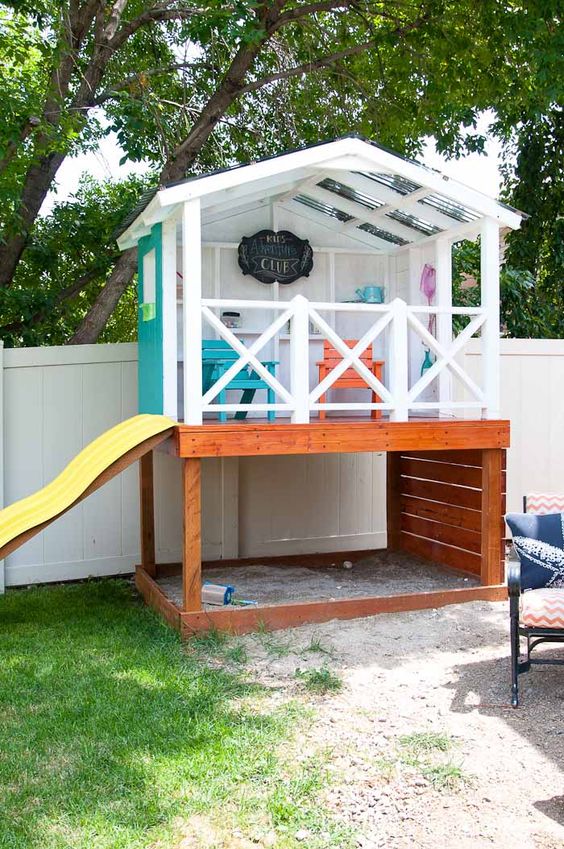 an outdoor playhouse with a yellow slide, with colorful furniture and some accessories and stained wood on one side