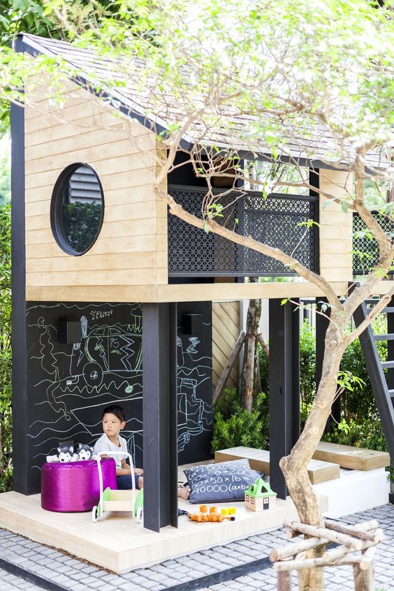 an ultra-modern two-level kids' playhouse with an upper part with a round window, a lower game spot with toys and low seats and a chalkboard