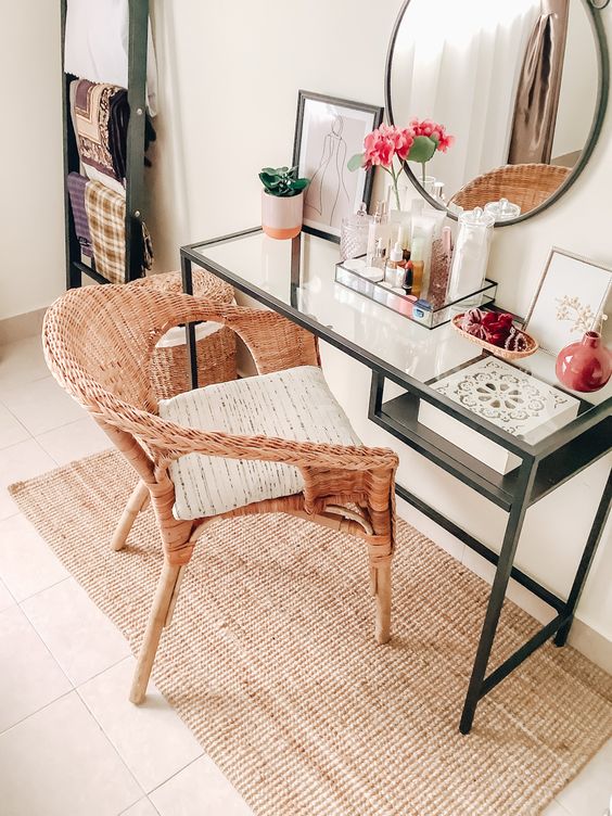 a chic vanity space with a Vittsjo desk, a round mirror, various decor makeup and a pink vase, a woven chair and a rug