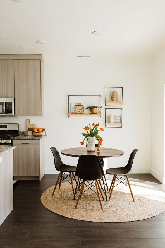 a small and lovely dining nook with a round table, black chairs, a woven rug and some open shelves with decor