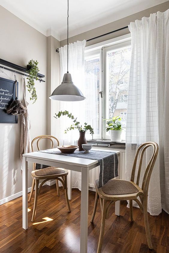 a small vintage dining nook with a white table, light stained chairs, a pendant lamp, greenery and a cute view