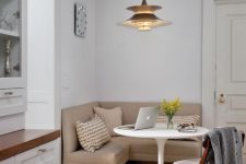 a stylish small dining nook with a tan corner bench, a round table, a white chair, a chic pendant lamp, printed pillows