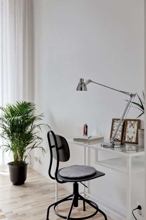 a stylish working space with a white Vittsjo desk, a black chair with a cushion, a silver table lamp and some art is elegant