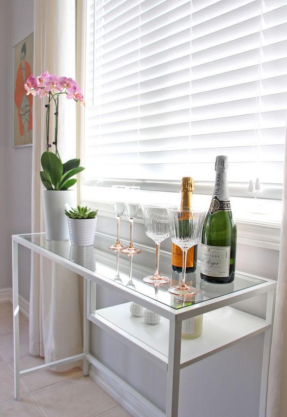 an elegant console table and home bar of a white Vittsjo desk, potted plants and blooms, elegant glasses and wine and champagne bottles