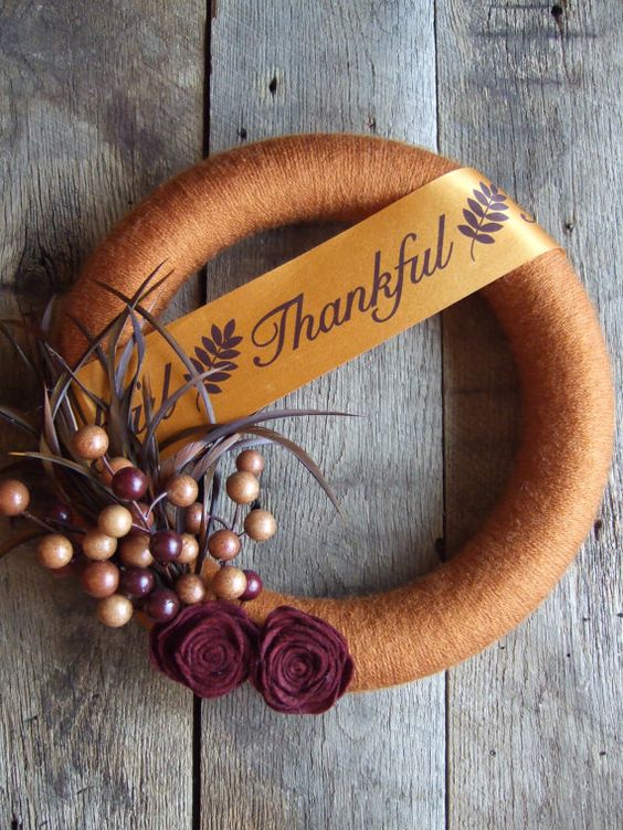 a Thanksgiving wreath covered with rust yarn, with fabric flowers, berries and feathers and a wide ribbon