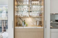 a chic built-in home bar with a marble backsplash, a mirror, glass shelves and sleek drawers for a modern space