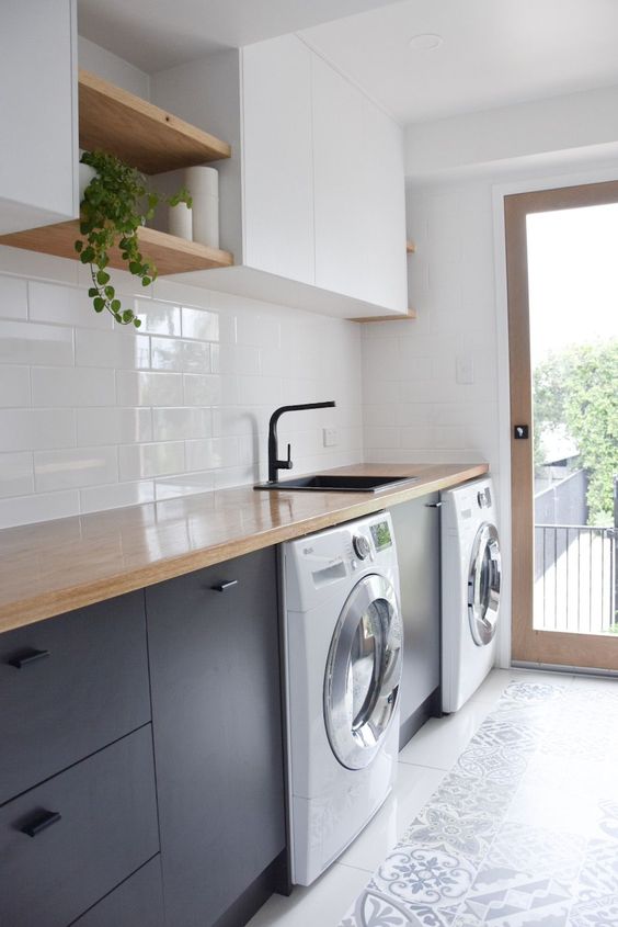 a chic modern laundry with sleke white and navy cabinets, butcherblock countertops, a washing machine and a dryer, a black faucet