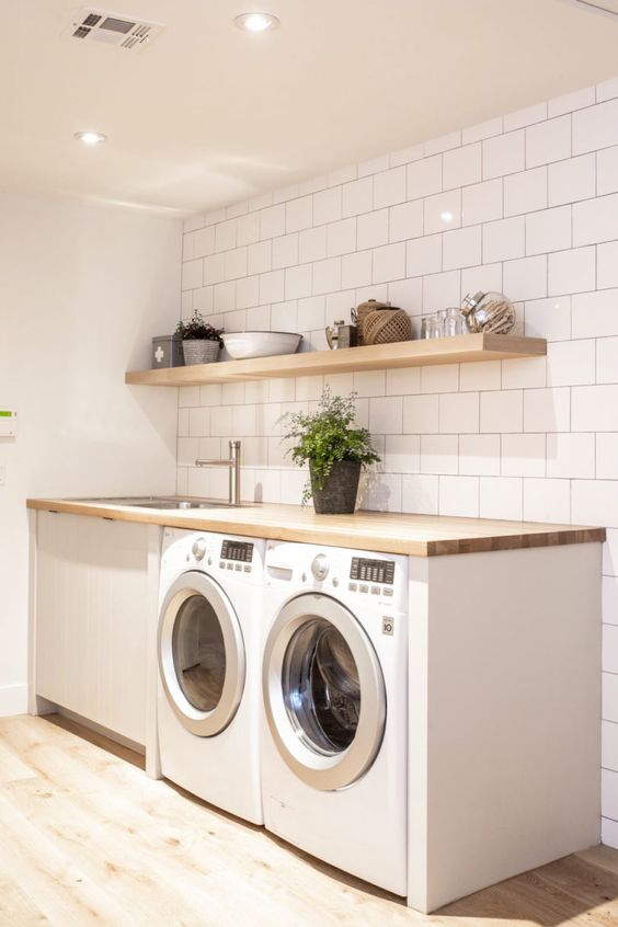 a clean white laundry with square tiles, an open shelf, a washing machine and a dryer, a butcherblock countertop and a potted plant