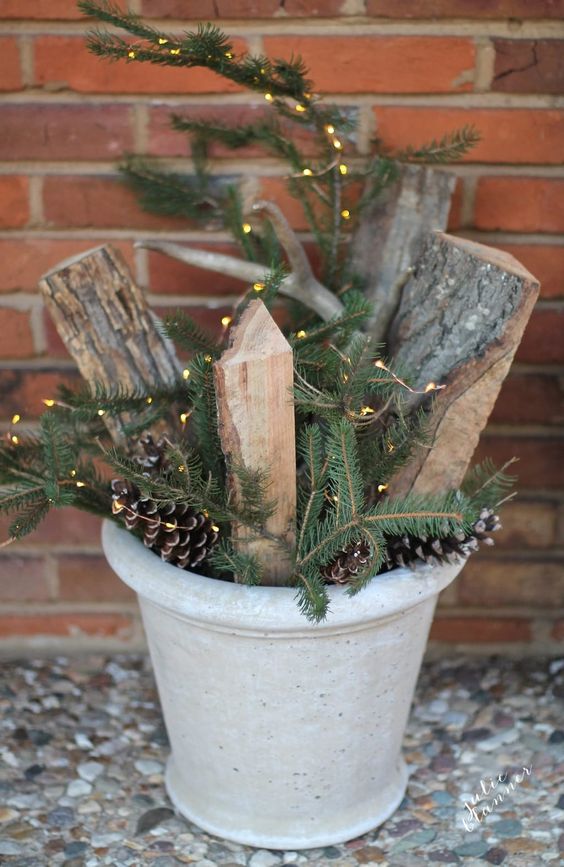 a concrete planter with pinecones, fir branches, firewood and lights is a cool outdoor decoration