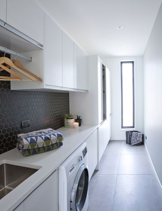 a contemporary laundry with sleek white cabinets, black hexagon tiles on the backsplash, a washing machine and a sink, a narrow window