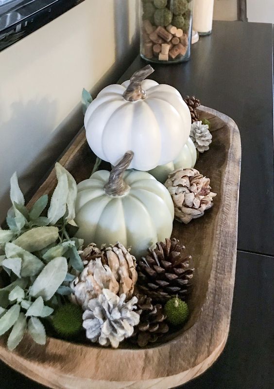 a fall decoration of a wooden bowl with pinecones, white faux pumpkins, green balls and pale leaves