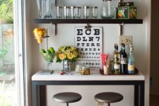 a home bar space with dark open shelves, a bar console, metal stools, blooms and an artwork make up a small yet chic nook