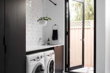 a laconic modern laundry room with sleek black cabinets, white tiles, a washing machine and a dryer and an entrace to the garden