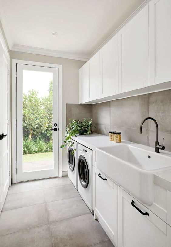 a laconic modern laundry with a concrete floor and walls, white shaker style cabinets, black handles and a door to the garden