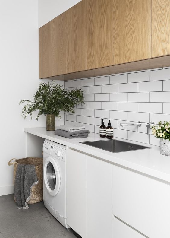 a modern laundry with sleek white and plywood cabinets, white countertops, white subway tiles and a washing machine plus a metal sink