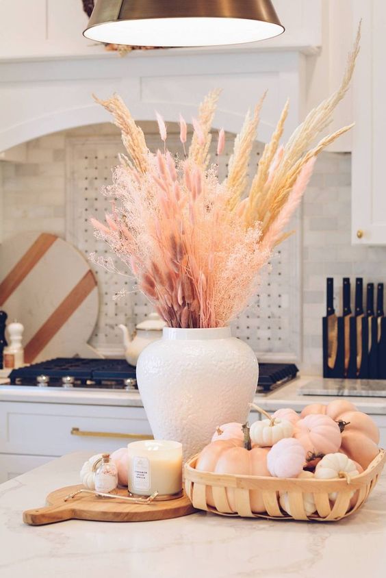 a pastel fall centerpiece of a pile of pumpkins in a wooden tray, a candle and some dried plants in a vase