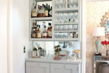 a stylish built-in home bar in dove grey, with a mirror backsplash, open storage compartments and a cabinet plus plants and blooms