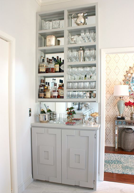 a stylish built-in home bar in dove grey, with a mirror backsplash, open storage compartments and a cabinet plus plants and blooms