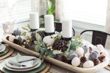 a stylish fall centerpiece of a white bowl with pinecones and mini pumpkins and pillar candles in the center