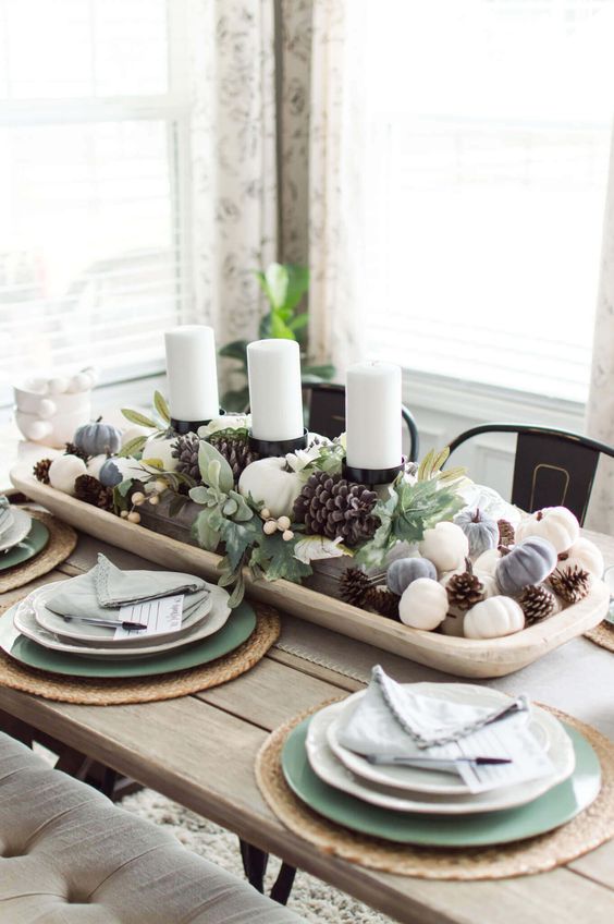 a stylish fall centerpiece of a white bowl with pinecones and mini pumpkins and pillar candles in the center