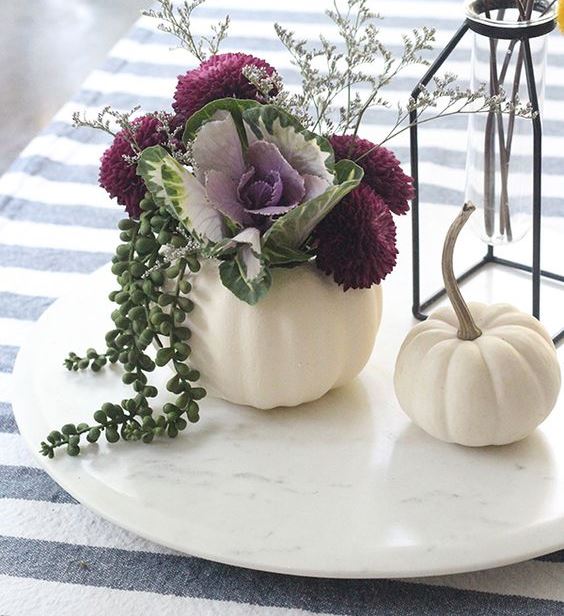 a white pumpkin with deep purple blooms, greenery, a cabbage, grasses and billy balls in a vase