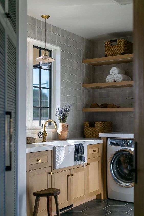 an elegant and welcoming laundry room with grey tiles, open shelves, stained cabinets, stone countertops, pendant lamps and a washing machine and a dryer