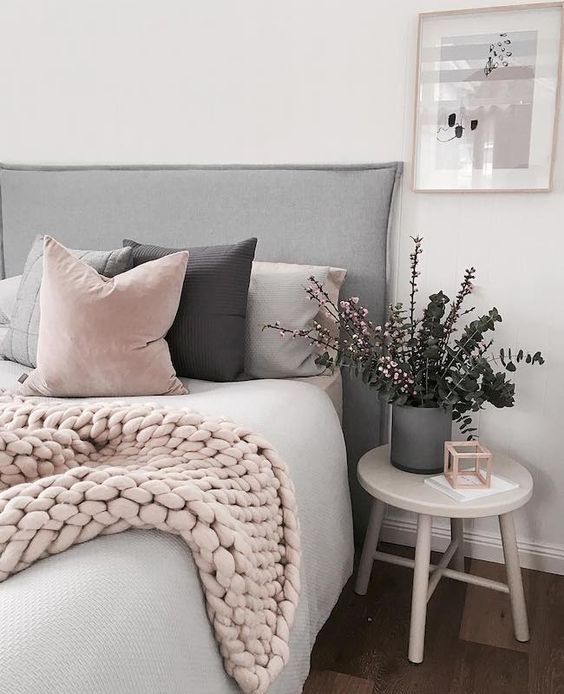 a Scandinavian bedroom with a grey upholstered bed, pink and grey pillows and a pink knit blanket, a grey planter with greenery