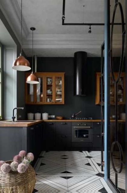 a black kitchen with matte cabinets, rich-stained upper ones, a black hood, copper pendant lamps and a black and white tile floor