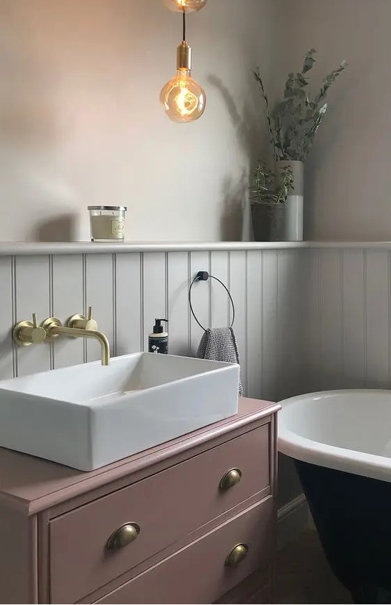 a chic bathroom with grey walls and white paneling, a black bathtub, a pink vanity, gold and brass fixtures