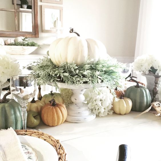 a chic fall centerpiece of a white pumpkin with lots of greenery on a white stand and faux pumpkins on the table