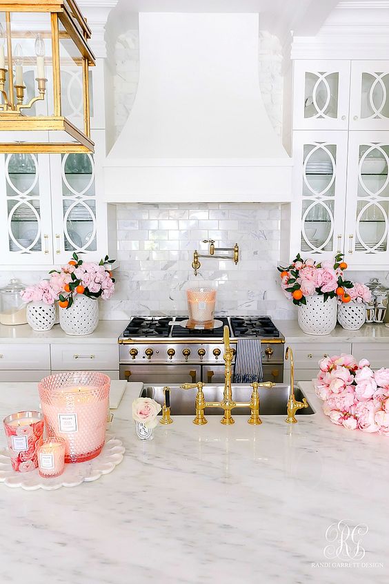 a chic glam kitchen with white cabinets, marble countertops, pink blooms, pink glasses with candles and gold touches
